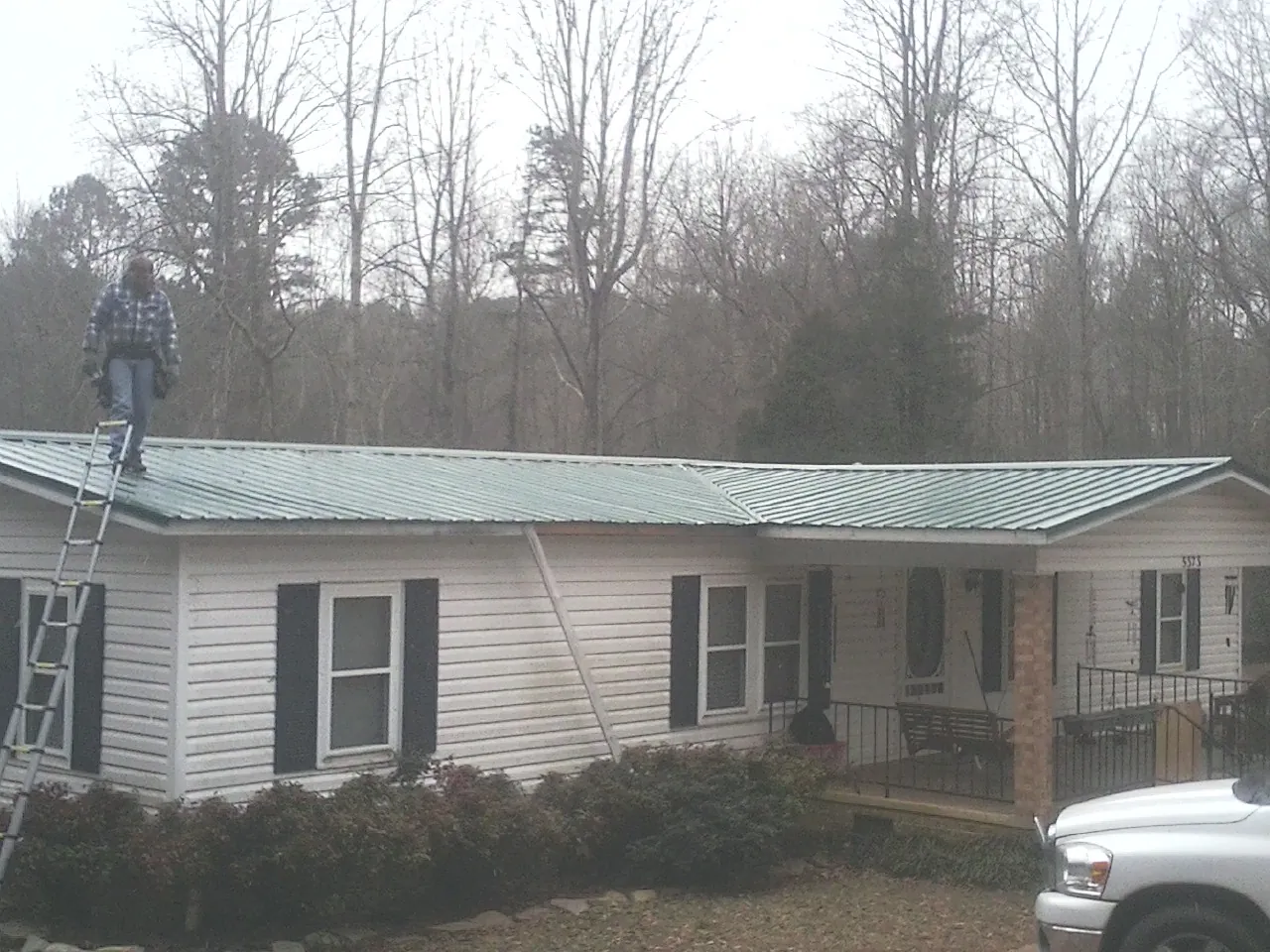 Worker on a ladder beside a new green metal roof on a ranch home in Magnolia